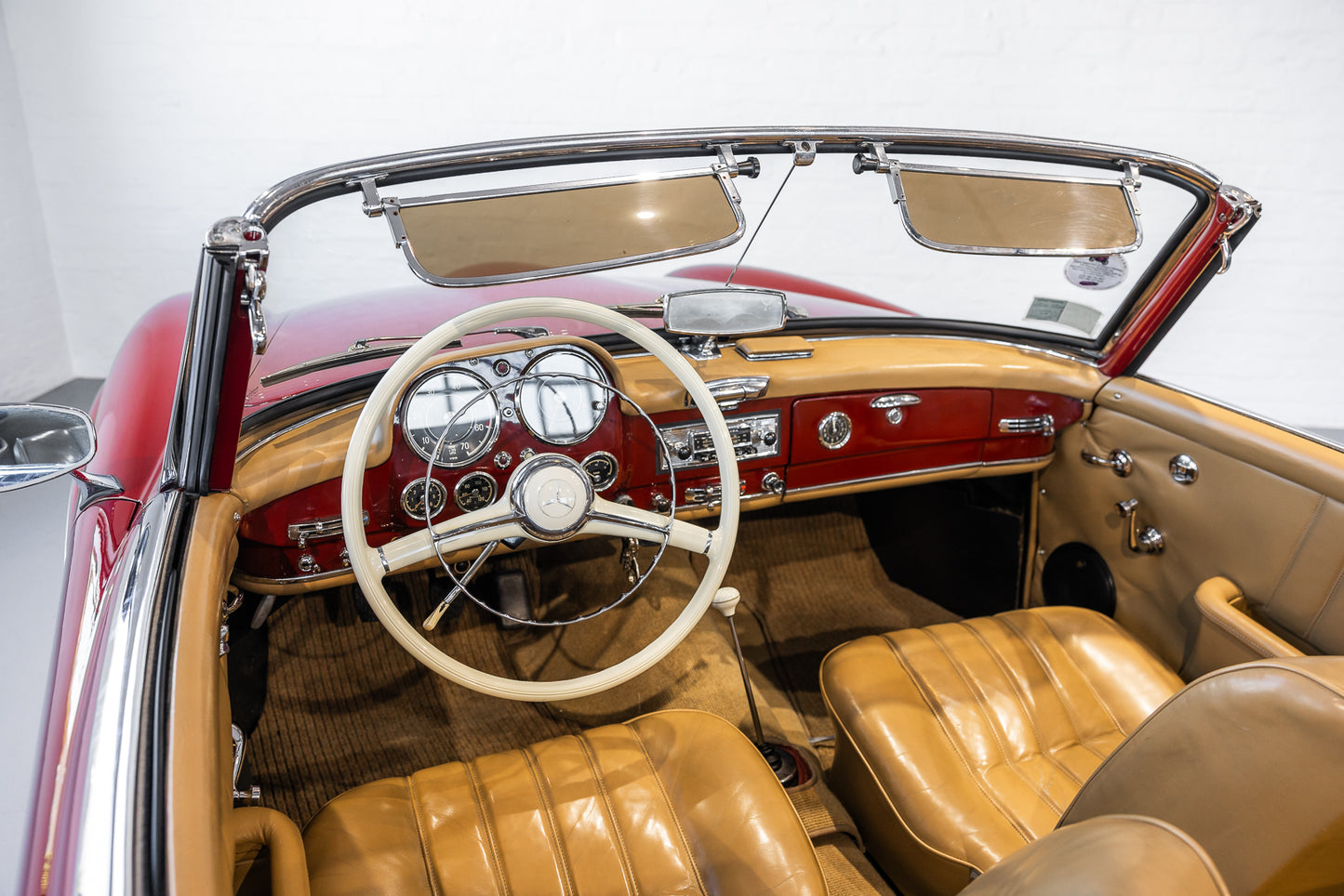 A vintage convertible car interior with tan leather seats and matching headrests, a cream steering wheel, and a red dashboard, photographed in a bright indoor setting. - Modern Classic Collective 190SL 