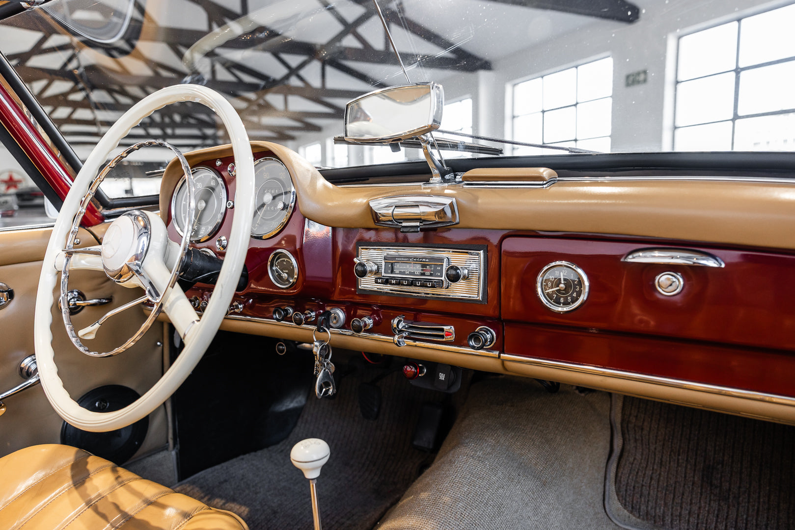 A close-up view of a classic car interior featuring a tan leather steering wheel, matching leather seats, a glossy dark red wood dashboard, chrome instrument gauges, and a vintage radio.